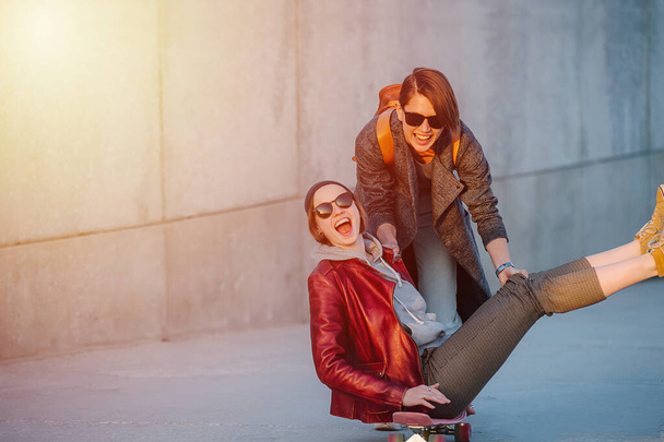 Portrait of two young amused women with short hair in sunglasses. One of them sitting on a skateboard with her legs in the air. Second one is giving a push to gain some speed. - Foto, immagini