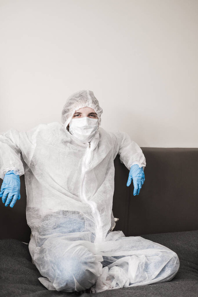 Two person in protective white translucent suit, blue rubber gloves, medical mask are sitting on sofa at home. Stay home during the coronavirus pandemic. - Photo, image