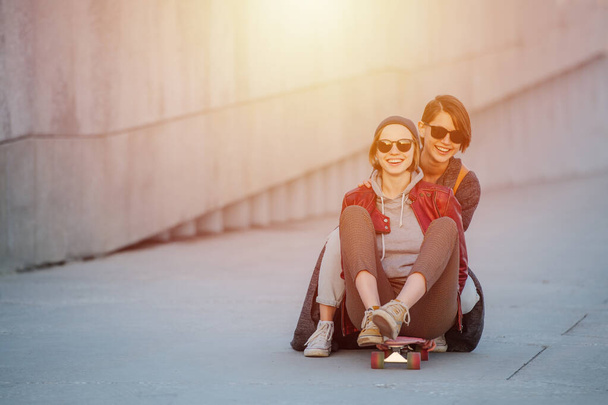 Retrato de duas jovens mulheres divertidas com cabelo curto em óculos de sol. Eles estão sentados em um skate, montando-o juntos
. - Foto, Imagem