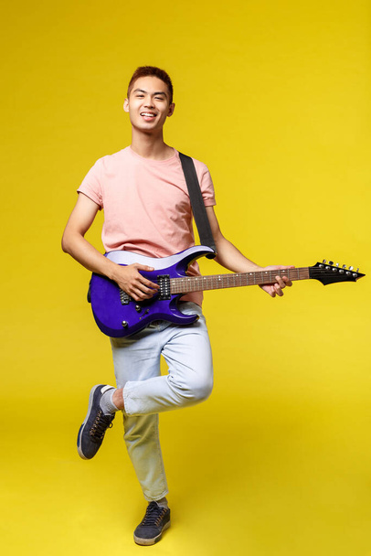 Életmód, szabadidő és ifjúság koncepció. Függőleges portré lelkes jóképű tinédzser ázsiai srác rózsaszín pólóban, játszik elektromos gitár, mosolygós boldog, álló sárga háttér - Fotó, kép