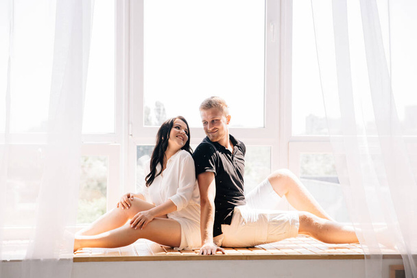 ζευγάρι κάθεται στο παράθυρο αγκαλιά και γέλιο και κοντά τους ένα λευκό τούλι σκορπίζει - Φωτογραφία, εικόνα