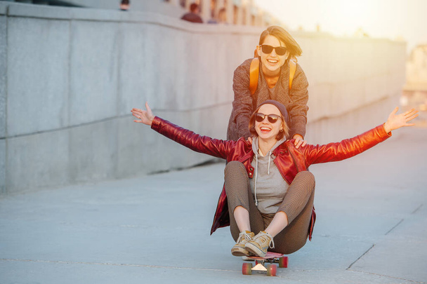 サングラスで短い髪を持つ2人の若い面白い女性の肖像画。そのうちの1人は、空中に彼女の足でスケートボードに座っています。二つ目は速度を上げるために彼女を押している. - 写真・画像