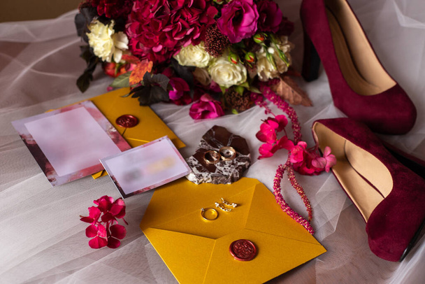 accesorios de boda: un ramo de novias de rosas, peonías y hortensias, anillos de boda en una piedra, un anillo y pendientes con diamantes, zapatos de mujer, invitación y sobres amarillos con un sello de cera roja
 - Foto, Imagen