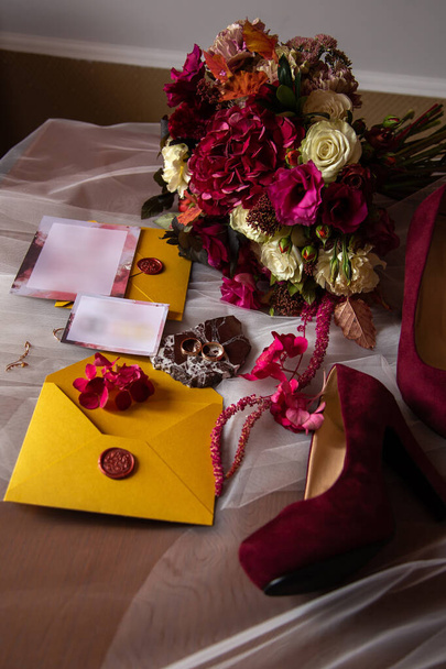 accesorios de boda: un ramo de novias de rosas, peonías y hortensias, anillos de boda en una piedra, un anillo y pendientes con diamantes, zapatos de mujer, invitación y sobres amarillos con un sello de cera roja
 - Foto, Imagen