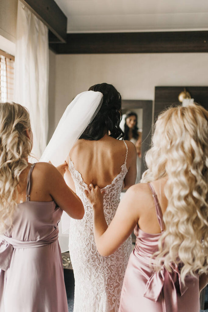 Bridesmaid helping slender bride lacing her wedding white dress, buttoning on delicate lace pattern with fluffy skirt on waist. Morning bridal preparation details newlyweds. Wedding day moments, wear. - Photo, Image