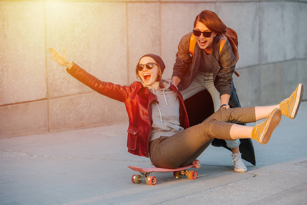 Portrait of two young amused women with short hair in sunglasses. One of them sitting on a skateboard with her legs in the air. Second one is giving a push to gain some speed. - Foto, Bild