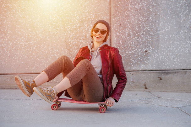 Portrait of a young amused woman with short hair in a red jacket, tight breeches and sunglasses. She's sitting on a skateboard with her legs in the air. - Fotoğraf, Görsel