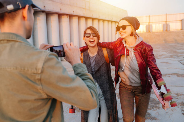 Man taking pictures of two young happy smiling women with short hair in sunglasses with skateboards. Sunset in a city. Gray floor and building. - Foto, Bild