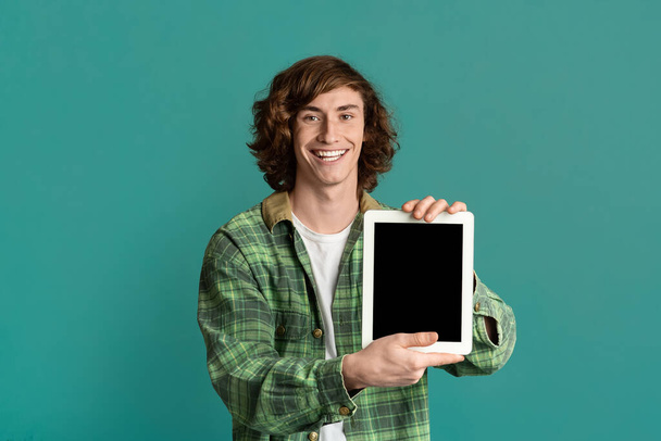Radosny nastolatek pokazujący tablet z pustym ekranem na turkusowym tle, przestrzeń do projektowania - Zdjęcie, obraz