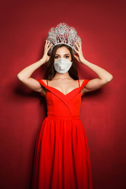 Όμορφη γυναίκα φορώντας ιατρική μάσκα προσώπου και διαμαντένιο στέμμα σε έντονο κόκκινο φόντο - Φωτογραφία, εικόνα
