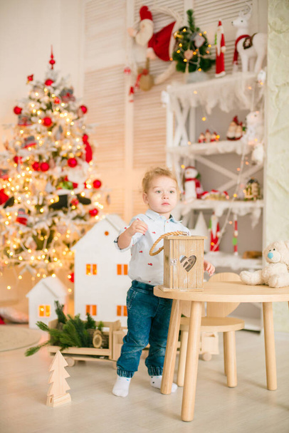 Cute little boy with blond hair plays with wooden eco toys in a bright room decorated with Christmas garlands near the Christmas tree. Happy childhood. Christmas mood - Zdjęcie, obraz