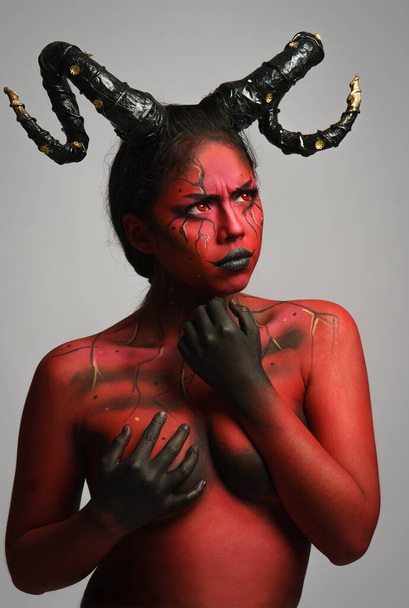 young pretty girl doing body painting characterized by demon evil with big black horns and red and black body doing poses in photo studio - Photo, Image