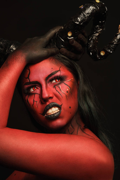 młoda ładna dziewczyna robi malowanie ciała charakteryzuje demona zła z dużymi czarnymi rogami i czerwony i czarny ciało robi pozy w studio fotograficznym - Zdjęcie, obraz