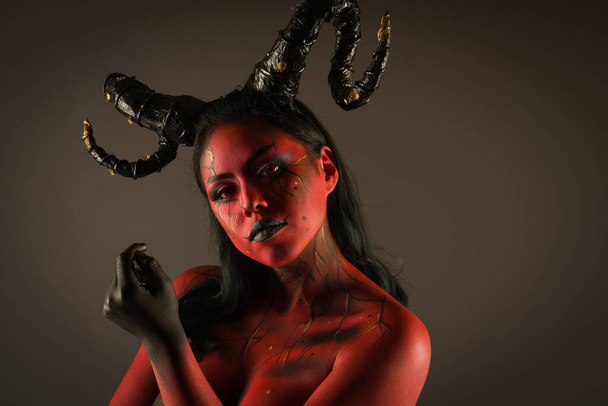 young pretty girl doing body painting characterized by demon evil with big black horns and red and black body doing poses in photo studio - Photo, Image