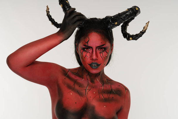 jong mooi meisje doen lichaam schilderen gekenmerkt door demon kwaad met grote zwarte hoorns en rood en zwart lichaam doet poses in fotostudio - Foto, afbeelding