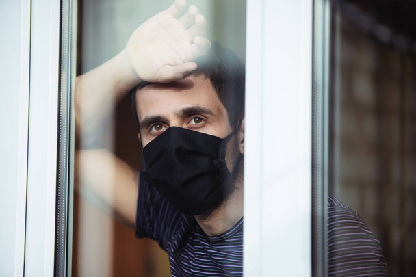 A man stays at home in isolation during quarantine. Coronavirus pandemic. The guy looks out the window into the street - Zdjęcie, obraz