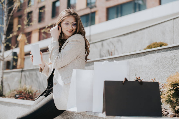 Aantrekkelijk schattig meisje met de boodschappentassen en warme drank in haar hand. Moderne stad megapolis op de achtergrond. Lady bestellen en online winkelen. Mooie Kaukasische dame - Foto, afbeelding