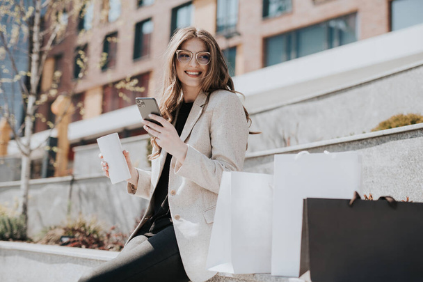 Attractive cute girl with the shopping bags and smarphone in her hand. Modern city megapolis on background. Lady making a phone call to order and shop online. Beautiful caucasian lady - Photo, image