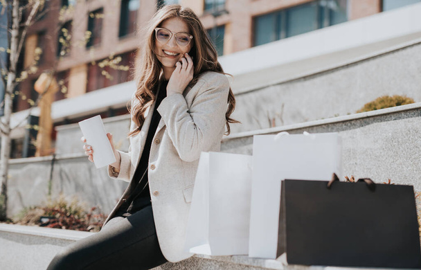Attraktives süßes Mädchen mit Einkaufstaschen und Smartphone in der Hand. Die moderne Megapolis der Stadt im Hintergrund. Lady telefoniert, um online zu bestellen und einzukaufen. Schöne kaukasische Dame - Foto, Bild