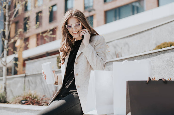 Attractive cute girl with the shopping bags and smarphone in her hand. Modern city megapolis on background. Lady making a phone call to order and shop online. Beautiful caucasian lady - 写真・画像