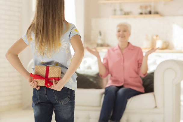 Little girl hiding present for grandmother behind her back, making surprise birthday gift at home. Blank space - 写真・画像