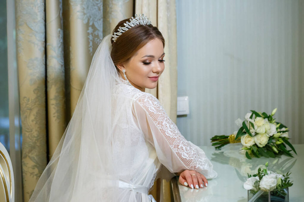 The bride in a white coat and veil sits at the makeup table - Fotografie, Obrázek