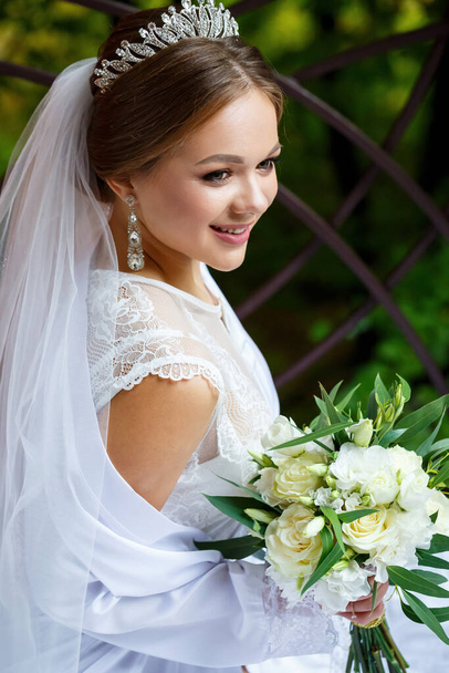 schöne Braut in Schleier und weißem Mantel sitzt auf einer Decke mit einem Brautstrauß in ihren Händen - Foto, Bild