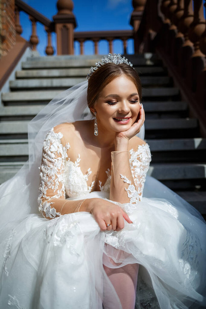 Gyönyörű menyasszony fehér esküvői ruhában ül a lépcsőn, és gyönyörűen mosolyog. - Fotó, kép
