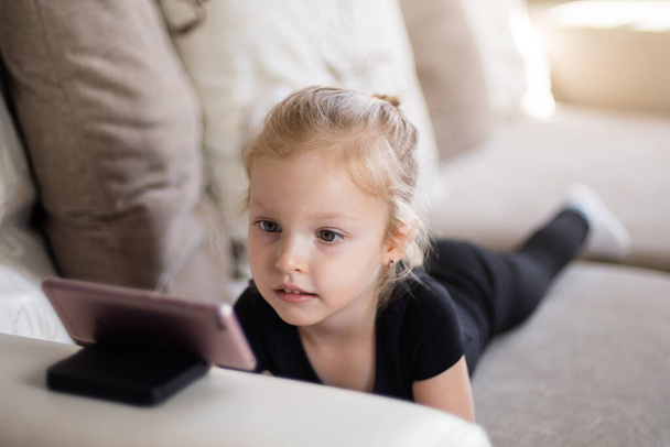 Etäopiskelua, verkko-opetusta lapsille. Tyttö opiskelee kotona älypuhelimen ääressä. Lapsi katselee piirrettyjä, lasten tietokoneriippuvuutta, vanhempien valvontaa. Karanteeni kotona - Valokuva, kuva