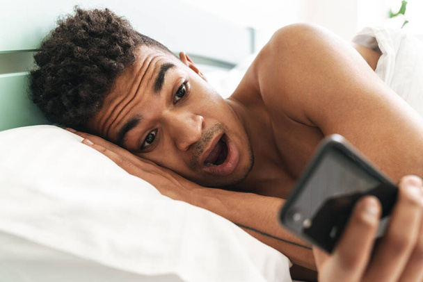 Image of shocked emotional young african guy in the morning in bedroom using mobile phone in bed. - Foto, Imagen