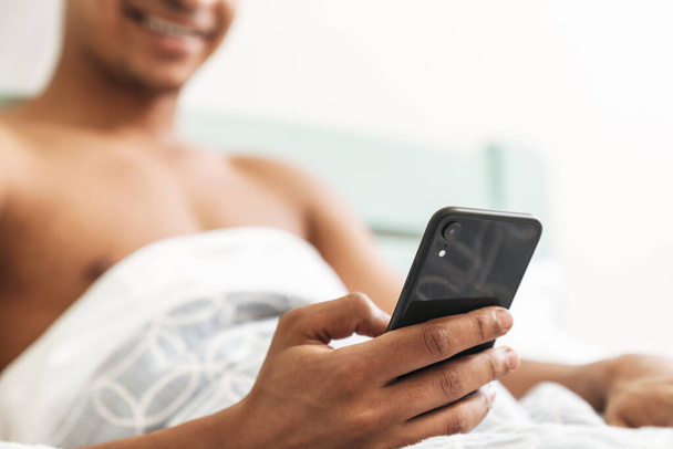 Cropped photo of optimistic pleased happy young african guy in the morning in bedroom using mobile phone in bed. - Foto, Imagem
