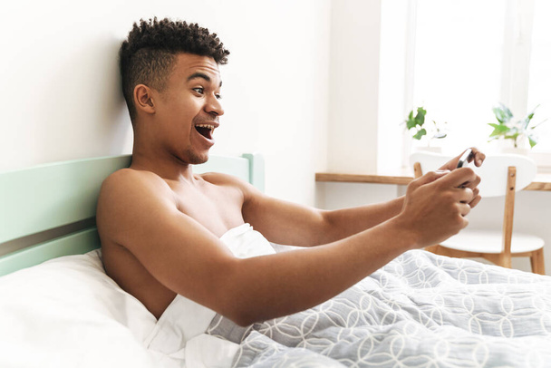 Image of emotional young african guy in the morning in bedroom play games by mobile phone in bed. - Fotoğraf, Görsel