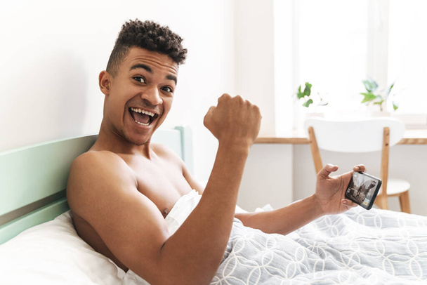 Image of happy positive young african guy in the morning in bedroom play games by mobile phone in bed make winner gesture. - 写真・画像