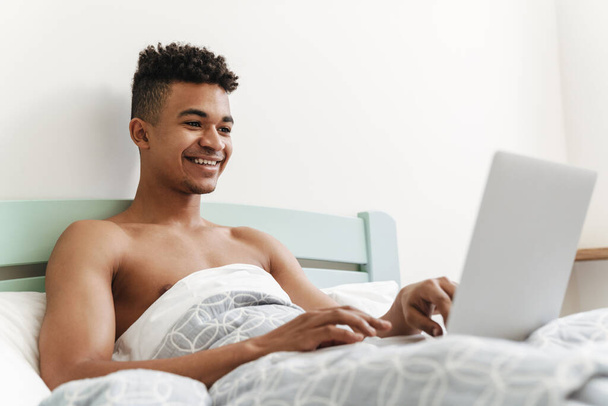 Photo of young happy positive african guy in the morning in bedroom using laptop computer in bed. - Фото, зображення