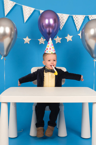 Happy blond boy is sitting at table in festive hat at birthday party, blowing festive whistle in joy and raising his hands to sides, celebrating achievement of goal, on decorated blue background. - Fotó, kép