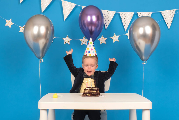 Happy caucasian boy is sitting at table in festive cone-shaped hat at birthday party, rejoicing and raising his hands to side, celebrating victory and achieving goal, on decorated blue background. - Fotó, kép