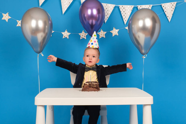 Happy caucasian boy is sitting at table in festive cone-shaped hat at birthday party, rejoicing and raising his hands to side, celebrating victory and achieving goal, on decorated blue background. - Φωτογραφία, εικόνα