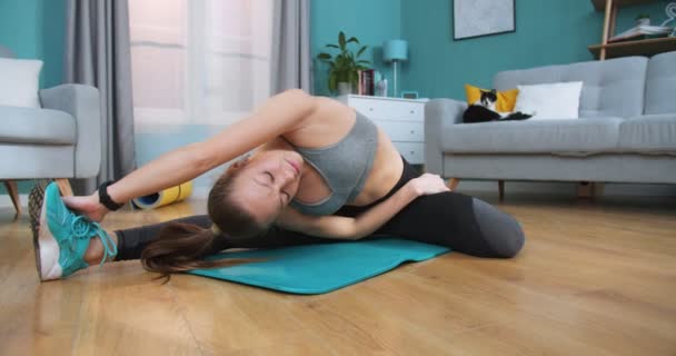 Female athlete in side position stretching on a blue mat on living room floor while the cat is sleeping in background. Sport and fitness. Training, workout concept. Active stretch girl doing sports. - Video