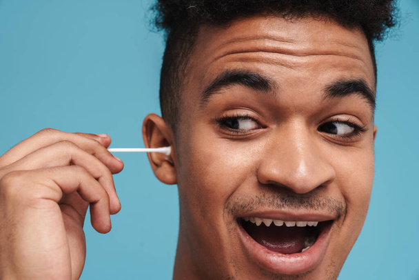 Photo of excited african american man using cotton swab and smiling isolated over blue background - Zdjęcie, obraz