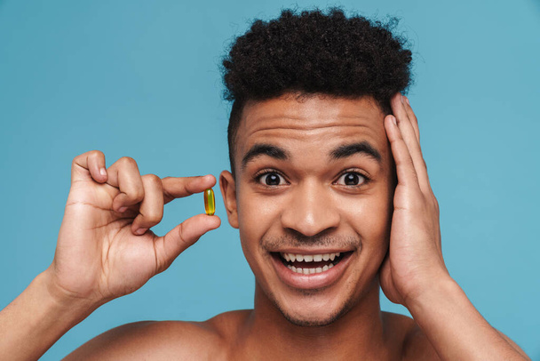 Photo of surprised african american man showing vitamin capsule isolated over blue background - Zdjęcie, obraz