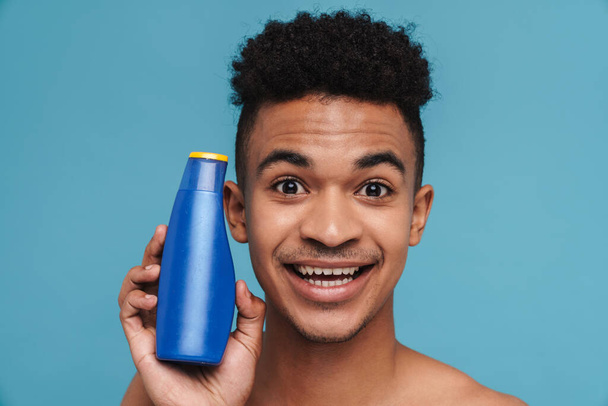 Photo of excited african american man showing shampoo and smiling isolated over blue background - Foto, Imagen