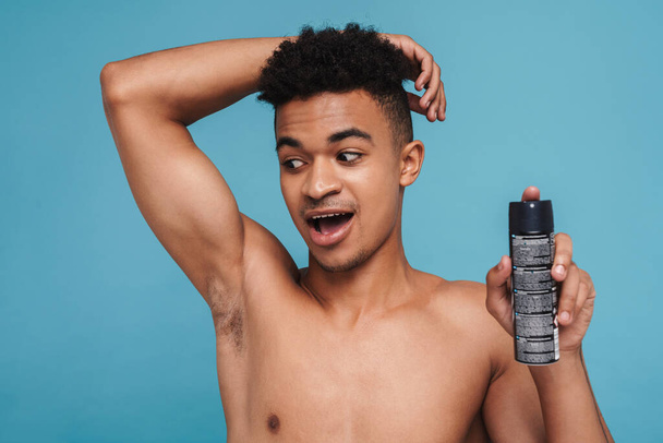 Photo of excited african american man showing his armpit and deodorant spray isolated over blue background - Photo, Image