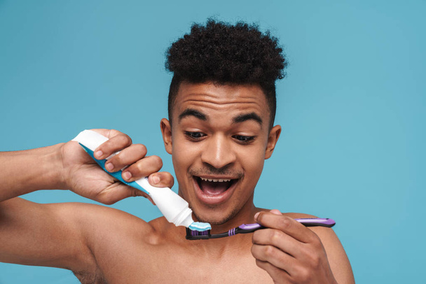 青い背景の上に隔離された歯磨き粉と歯ブラシを使用している間に微笑む白人のアフリカ系アメリカ人の男性の写真 - 写真・画像