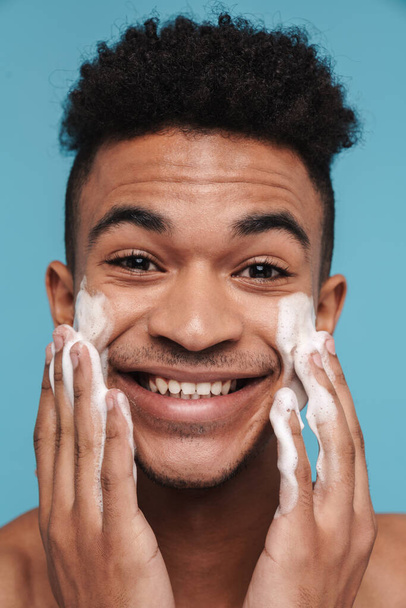 Photo of shirtless african american man smiling while applying facial cleanser foam isolated over blue background - Valokuva, kuva