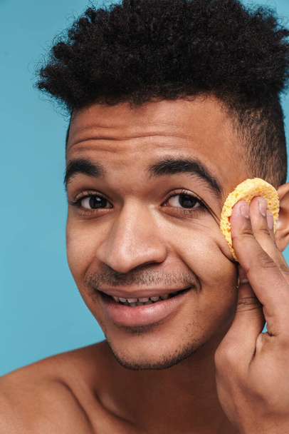 Photo of shirtless african american man smiling while cleaning his face with sponge isolated over blue background - Foto, imagen