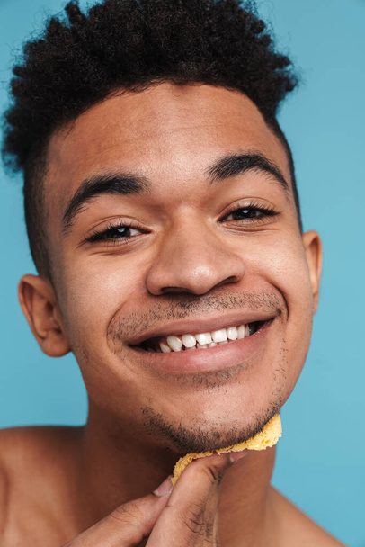 Photo of shirtless african american man smiling while cleaning his face with sponge isolated over blue background - Foto, Bild