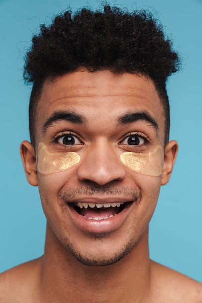 Фотографія збудженого афроамериканського чоловіка з під очними плямами посміхається в камері, ізольованій над синім фоном. - Фото, зображення