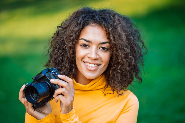 Young pretty african woman in yellow takes pictures with DSLR camera over green background. Girl smiling having fun as photographer. - Foto, immagini
