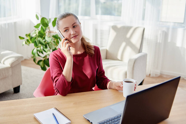 Happy young business woman sitting at wood desk, enjoying tea and talking on cellphone at home office, copy space - Фото, изображение