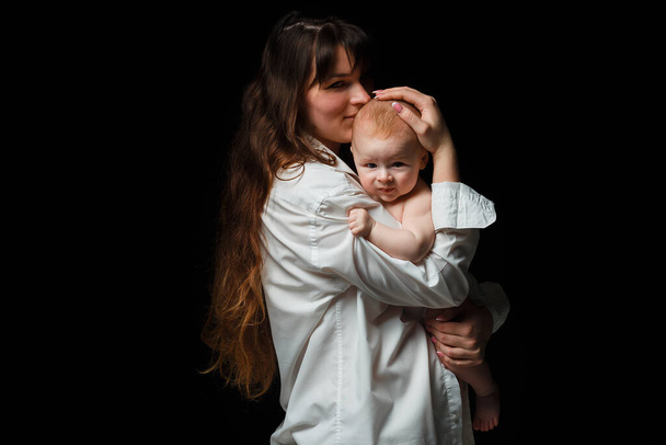 young brunette mom with long hair and a white shirt holds a newborn son. A 4 month old baby in her arms. On a black background. - Photo, Image